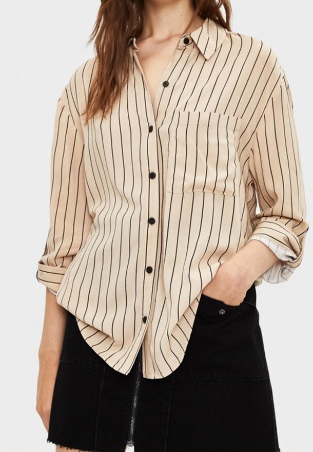 Femme portant une chemise beige à boutons avec des rayures verticales noires et une jupe noire avec une fermeture éclair devant, main dans la poche.