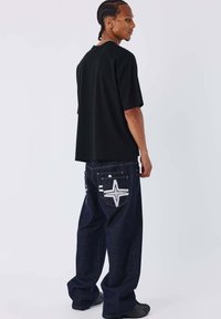 Maglietta nera a maniche corte abbinata a jeans di denim scuro larghi con disegni bianchi a stella e strisce sulle tasche posteriori.