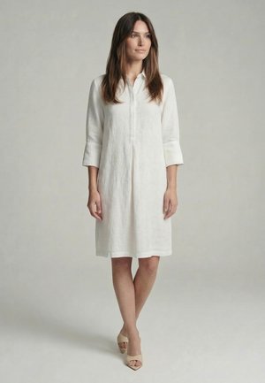 Woman standing in a white knee-length dress with three-quarter sleeves and beige open-toe heels, against a plain light gray background.