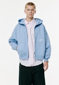 Sweat à capuche bleu clair avec fermeture éclair sur le devant, présentant une finition texturée, de grandes poches et un logo brodé sur la poitrine. Porté par-dessus une chemise rayée.