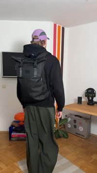 Sac à dos noir à rabat enroulable avec bretelles ajustables, doté d'une boucle avant et d'une poche latérale, porté avec un pantalon vert olive et un sweat à capuche noir.