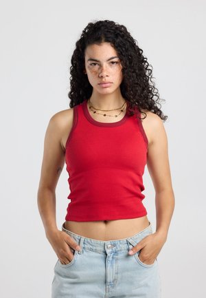 Vrouw met lang krullend haar, draagt een rood hemdje en lichtblauwe jeans, staat vooraan met haar handen in de zakken tegen een effen achtergrond.