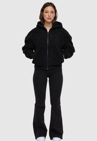 Black, textured fleece zip-up hoodie with a hood and side pockets, paired with black flared jeans and white sneakers.