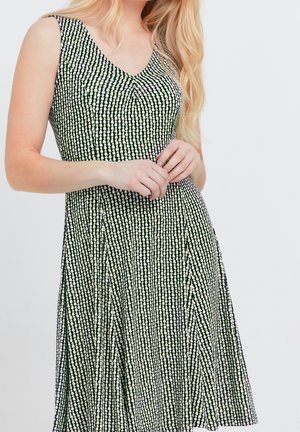 Mujer vestida con un vestido sin mangas de patrón blanco y negro con escote en V, con las manos suavemente entrelazadas en la cintura y el cabello largo y rubio sobre el hombro.