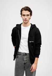 Joven con sudadera con cremallera negra sobre una camiseta blanca y jeans lavados grises, de pie frente a un fondo claro y sencillo.