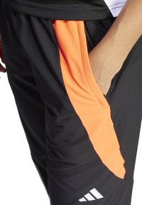 Schwarze Sporthosen mit orangefarbenen Seitenpanelen, ausgestattet mit einer Reißverschlusstasche und einem strukturierten Stoff. Enthält ein Logo in Weiß nahe dem Saum.