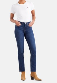 Jeans de mezclilla de tiro alto en un color azul oscuro, combinados con una camiseta blanca ajustada que presenta un pequeño logo rojo, y botas de tobillo color beige con tacones anchos.