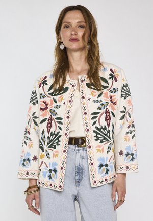 Femme portant une veste crème avec des motifs floraux brodés colorés, un jean clair et des boucles d'oreilles créoles dorées sur un fond uni.