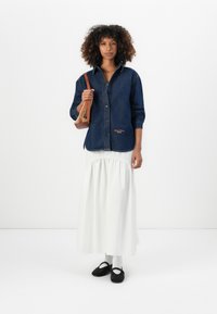 Vrouw met krullend haar, gekleed in een donkerdenim overhemd, lange witte rok, zwarte schoenen met witte sokken en een bruine schoudertas.