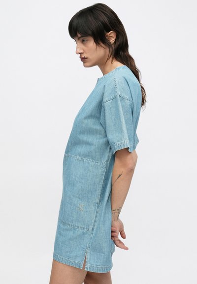 Γυναίκα με κοντομάνικο ανοιχτό μπλε φόρεμα από τζιν με πλευρικές σκισμές, μαλλιά μαζεμένα πίσω και τατουάζ στο αριστερό αντιβράχιο, σε λευκό φόντο.