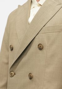 Μπεζ σακάκι διπλής σειράς κατασκευασμένο από υφασμάτινο υλικό με υφή, διαθέτοντας μεγάλους καφέ κουμπιά και μια μόνο τσέπη στο στήθος. Μαλακό σχέδιο γιακα.