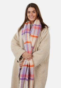 Bufanda a cuadros en una suave mezcla de lana con tonos naranja, púrpura y pastel; con bordes flecados; envuelta alrededor del portador que lleva un abrigo beige.