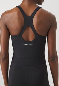 Melnā sporta toppenes ar sacensību aizmuguri un izgriezumu. Satur logo "SWEATY BETTY" baltā krāsā, gludu tekstūru un pieguļošu siluetu.