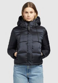 Giacca puffer isolata blu navy con cappuccio staccabile, chiusura frontale con zip, cordini elastici e tasche laterali. Texture liscia e lucida.