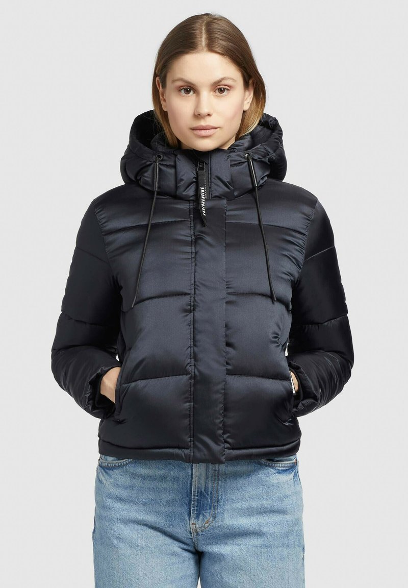 Giacca puffer isolata blu navy con cappuccio staccabile, chiusura frontale con zip, cordini elastici e tasche laterali. Texture liscia e lucida.