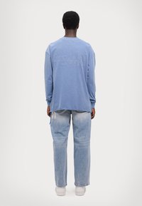 Felpa a maniche lunghe color azzurro chiaro con logo "KSUBI" in rilievo sulla schiena, abbinata a jeans in denim chiaro a vestibilità comoda e sneakers bianche.