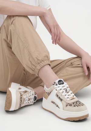Persona seduta con pantaloni beige e sneakers bianche platform con stampa leopardata e lacci bianchi su sfondo bianco.