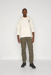 Kremasta jopica s kapuco in grafičnim potiskom, kombinirana z olivno zelenimi cargo hlačami z žepki z zadrgo ter črnimi pohodnimi čevlji.