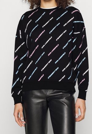 Personne portant un sweat à capuche noir avec le texte "KARL LAGERFELD" en diagonale en bleu, blanc et rose, associé à un pantalon en cuir noir.