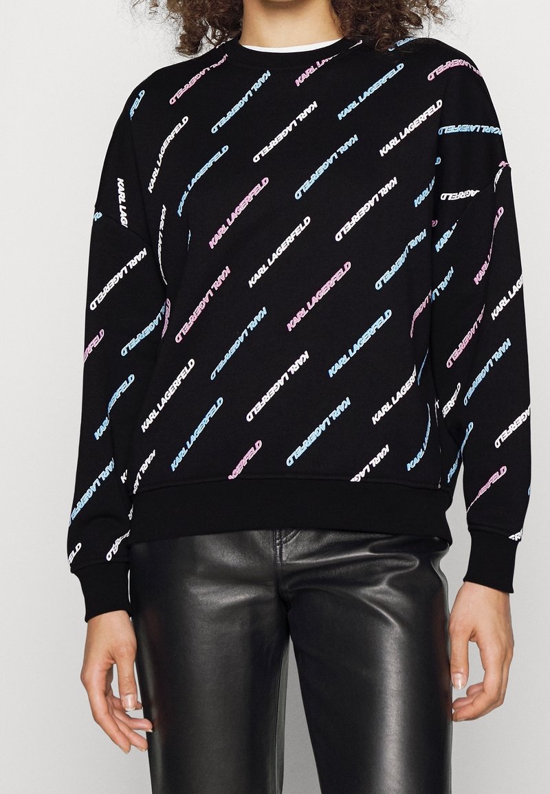Persona che indossa una felpa nera con la scritta diagonale "KARL LAGERFELD" in blu, bianco e rosa, abbinata a pantaloni di pelle neri.