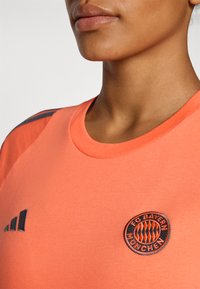FC Bayern München oranje sportshirt met een ronde hals, voorzien van een zwart geborduurd embleem en drie zwarte accentstrepen op de schouder.