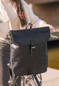 Zaino in pelle nera con pattina pieghevole, tracolla regolabile e chiusura a fibbia. Presenta una texture liscia e un design minimalista.