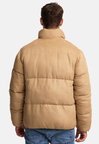 Beżowa kurtka puchowa z wysokim kołnierzem, pikowanym wzorem i luźnym krojem. Posiada poziome przeszycia oraz gładkie, lekko błyszczące wykończenie.