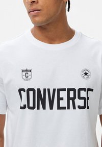 Camiseta de algodón blanca con texto en negrita negro "CONVERSE", un pequeño logo en el lado izquierdo del pecho y un logo redondo en el hombro derecho.