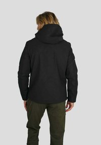 Personne aux cheveux mi-longs portant une veste noire à capuche et un pantalon vert olive, se tenant de dos face au spectateur contre un fond uni.