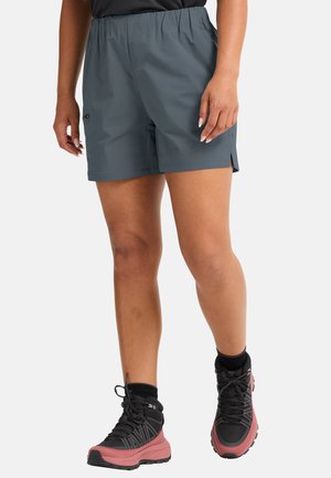 Personne portant un short de randonnée gris et des bottes de randonnée noires et roses, debout avec les mains détendues le long du corps sur un fond blanc.