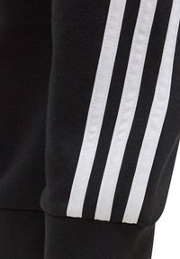 Pantalones deportivos negros con tres franjas verticales blancas a lo largo de las costuras laterales. Elaborados con una tela suave de textura lisa.