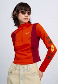 Nike langermet topp har oransje og maroon paneler, høy krage og logodetaljer i svart og gult på ermet. Glatt tekstur.