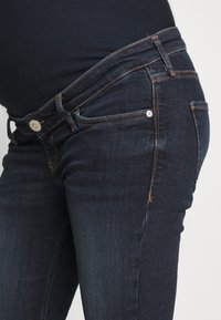 Primer plano de una barriga de embarazada que lleva jeans de maternidad oscuros con una cinturilla elástica y costuras naranjas.
