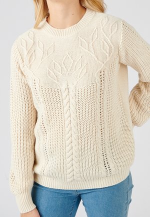 Pull en maille torsadée crème avec motifs de feuilles porté par une personne debout, une main sur la hanche, associé à un jean bleu.