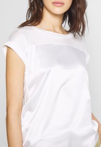 White satin short-sleeve top with a smooth texture, rounded neckline, and rolled cuffs. Features a subtle sheen and loose fit design.