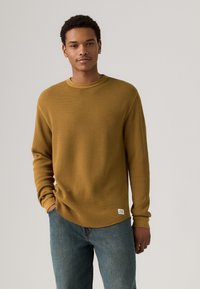 Sennep gul ribbet langermet skjorte med rund hals og oppbrettede ermer, kombinert med blå denimjeans. Merket synlig nederst til venstre.