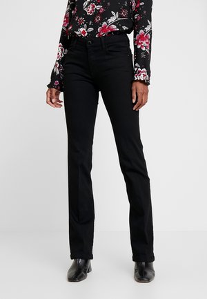 Person trägt eine schwarze, gerade geschnittene Jeans, eine schwarze geblümte Langarmbluse und schwarze Stiefeletten und steht vor einem schlichten Hintergrund.