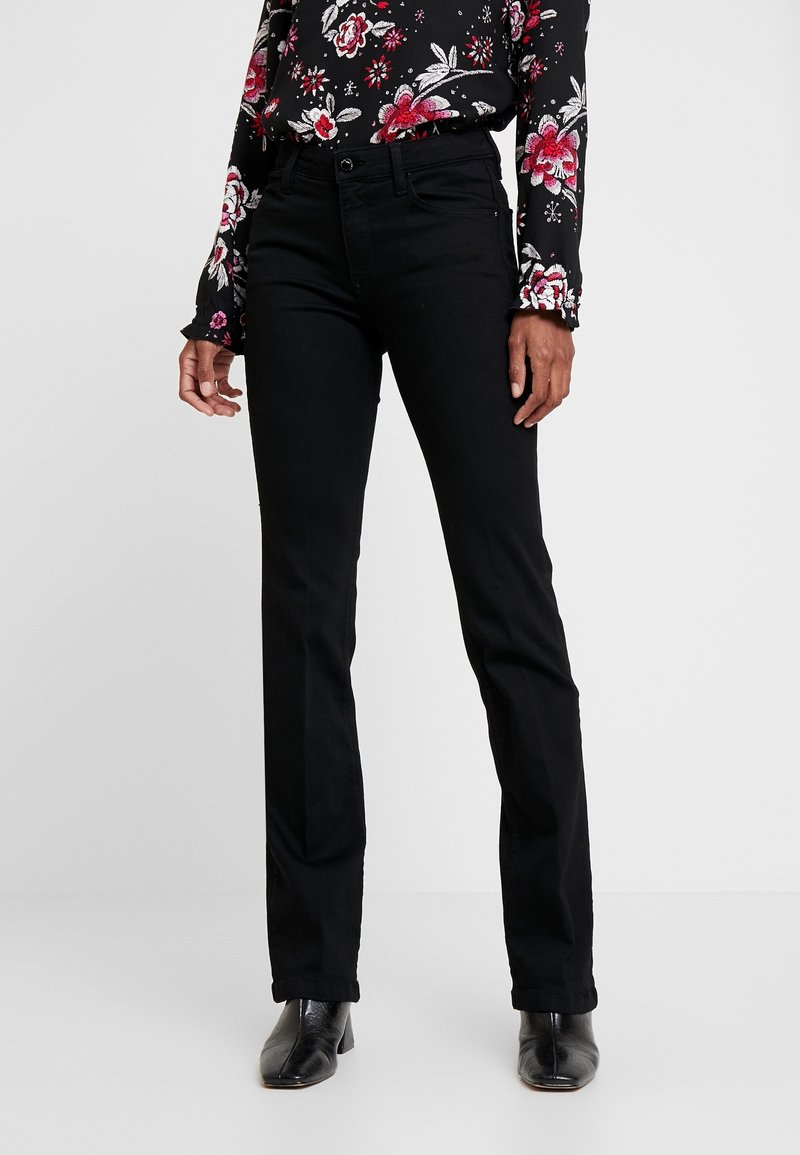 Personne portant un jean noir droit, un chemisier noir à manches longues à motif fleuri et des bottines noires, debout devant un fond uni.