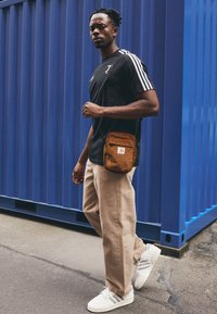 Sort atletisk t-shirt med hvide striber, beige baggy bukser og hvide sneakers med grå detaljer. Brun korduroy taske slynget over kroppen.
