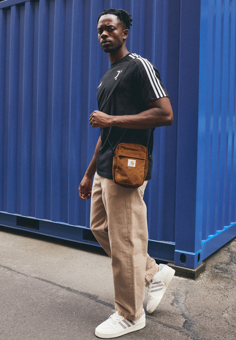 T-shirt de sport noir avec des rayures blanches, pantalon beige ample et baskets blanches avec des accents gris. Sac en velours côtelé brun porté en bandoulière.