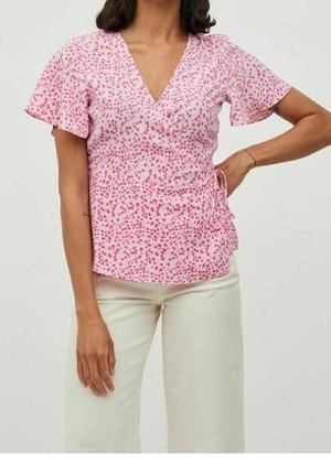 Nainen, joka pukeutunut vaaleanpunaiseen lyhythihaiseen kietaisupaitaan, jossa on pieni punainen kukkakuviointi, ja luonnonvalkoisiin leveälahkeisiin housuihin, seisoo käsi lanteilla.