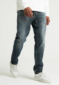 Blugi negri cu un design decolorat, având o tăietură dreaptă și o ușoară uzură. Purtați cu adidași albi și un top alb cu mânecă lungă.