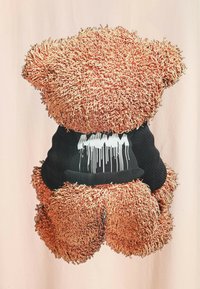 Osito de peluche con un cuerpo texturizado de color marrón claro y desaliñado, vistiendo una camisa negra con un diseño gráfico goteante en blanco. El fondo es claro.