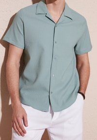 Koszula z krótkim rękawem w jasnym zielonym kolorze, wykonana z teksturowanej tkaniny z subtelnym prążkowanym wzorem, z kołnierzykiem w kształcie litery V oraz zapięciem na guziki.