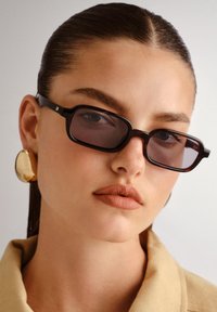 Ochelari rectangulari negri cu lentile întunecate, asociați cu un cercei sculptural din aur. Textură netedă pe un top bej.