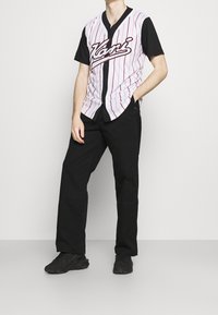 Maglia stile baseball con strisce verticali rosse su sfondo bianco, maniche nere e testo "Kani"; abbinata a pantaloni neri e sneakers.