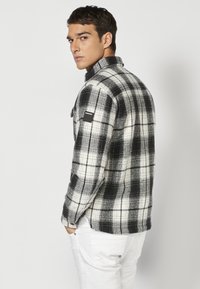 Zwart-wit geruite flanellen shirt met een relaxte pasvorm, kraag en een logo patch op de linker schouder. Het model draagt witte broeken.