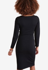 Robe noire à manches longues avec une texture côtelée, une silhouette ajustée et un col rond, présentant une coupe jusqu'au genou sans embellissements visibles.