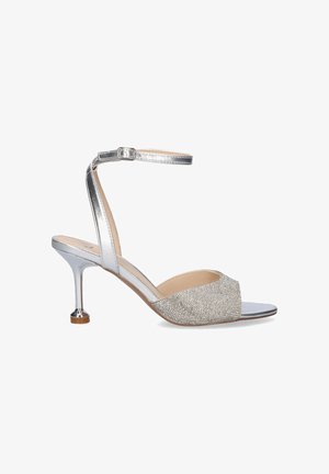 Sandalia plateada de tacón alto con correa en el tobillo y punta abierta, con una banda frontal texturizada y brillante y un tacón delgado y curvado.