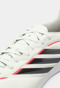 Κοντινό πλάνο ενός λευκού αθλητικού παπουτσιού με μαύρες και κόκκινες διαγώνιες ρίγες και λευκές κορδόνες σε φωτεινό φόντο.